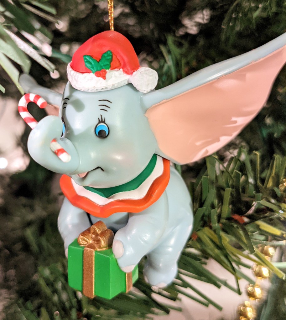 Christmas tree ornament - Dumbo, an elephant with very large ears, is wearing a red Santa hat with holly berry on it, is holding a red and white candy cane with his trunk, and is holding a green present with gold ribbon with his front feet. He looks very happy.