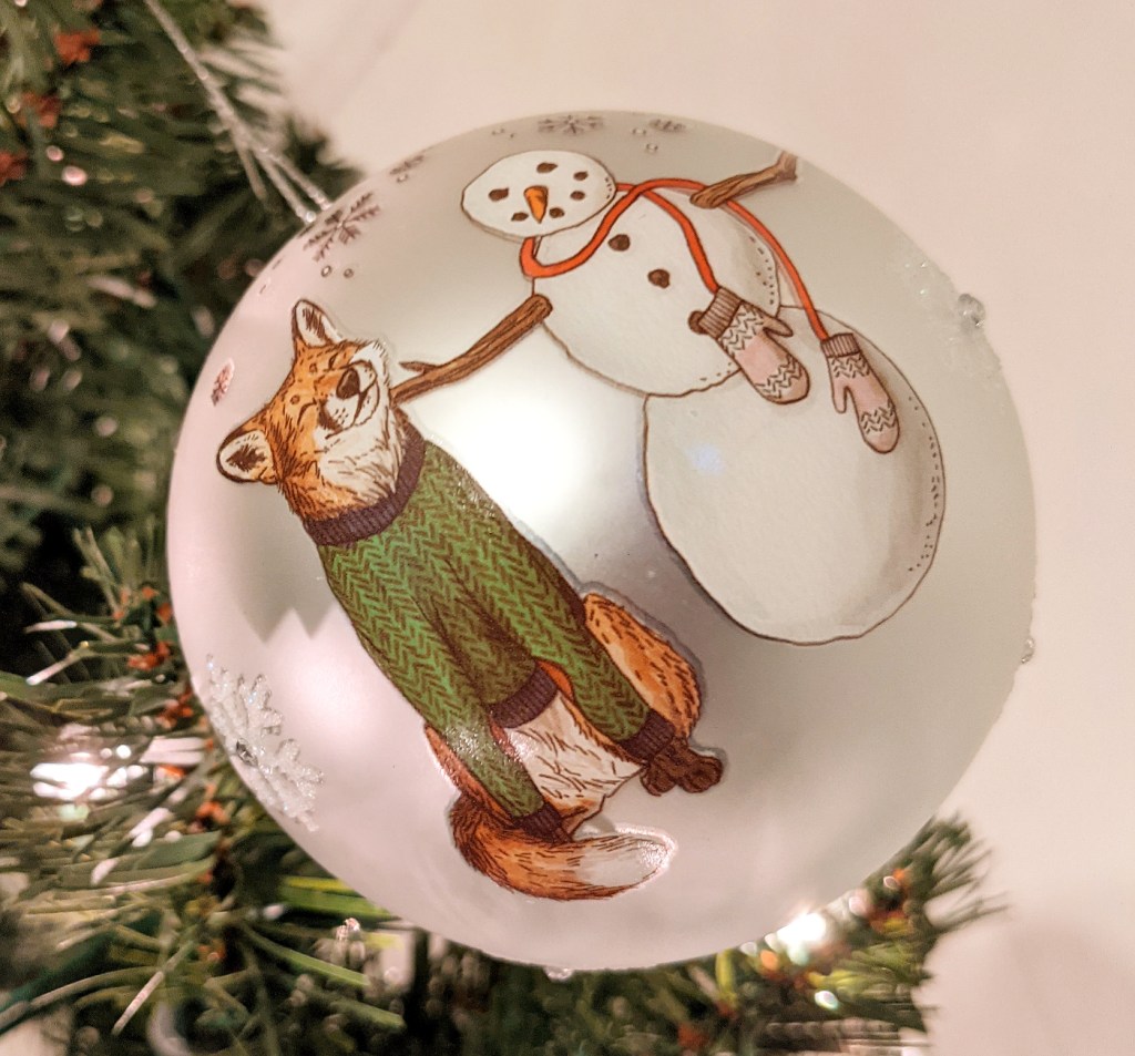 A red fox wearing a green sweater happily rubs its chin against a snowman. This image is on a silver ball that has sparkly snowflakes sporadically in the background.