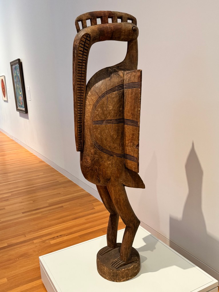 a wooden sculpture of a bird; the following was on a plaque next to the sculpture: 

Artist unrecorded
Senufo, Côte d'Ivoire
Sejen (bird)
Before 1966
Wood
Loan from Marlene Ross
Provenance: Sidney Kamerman 1966
This monumental sculpture was bought in 1966 by Sidney Kamerman while he was based in Yamoussoukro, the capital of Côte d'Ivoire, as a Peace Corps volunteer. Kamerman taught at a local vocational school, training young Ivorians to become car mechanics. He purchased the sculpture directly from the artist and transported it back to the United States-quite an ordeal at the time. Indeed, the current owner of the sculpture, Kamerman's sister and long-time UMMA docent Marlene Ross, recalls that the large crack running down the middle of the sculpture occurred during shipping, when the crate housing the figure was in the French port of Marseille, awaiting the approval of customs paperwork.
Bird figures were revered by the Senufo of Côte d'Ivoire, who displayed them only on important occasions, such as funerals and initiation celebrations. Sometimes they were worn on the head as demonstrations of the wearer's strength and virility. While such sculptures were originally very rare and only accessible to members of the Poro, a cross-regional society for initiated men, they have since become ubiquitous carvings for export, and can now be purchased at tourist markets all over the continent.
PLEASE DO NOT TOUCH