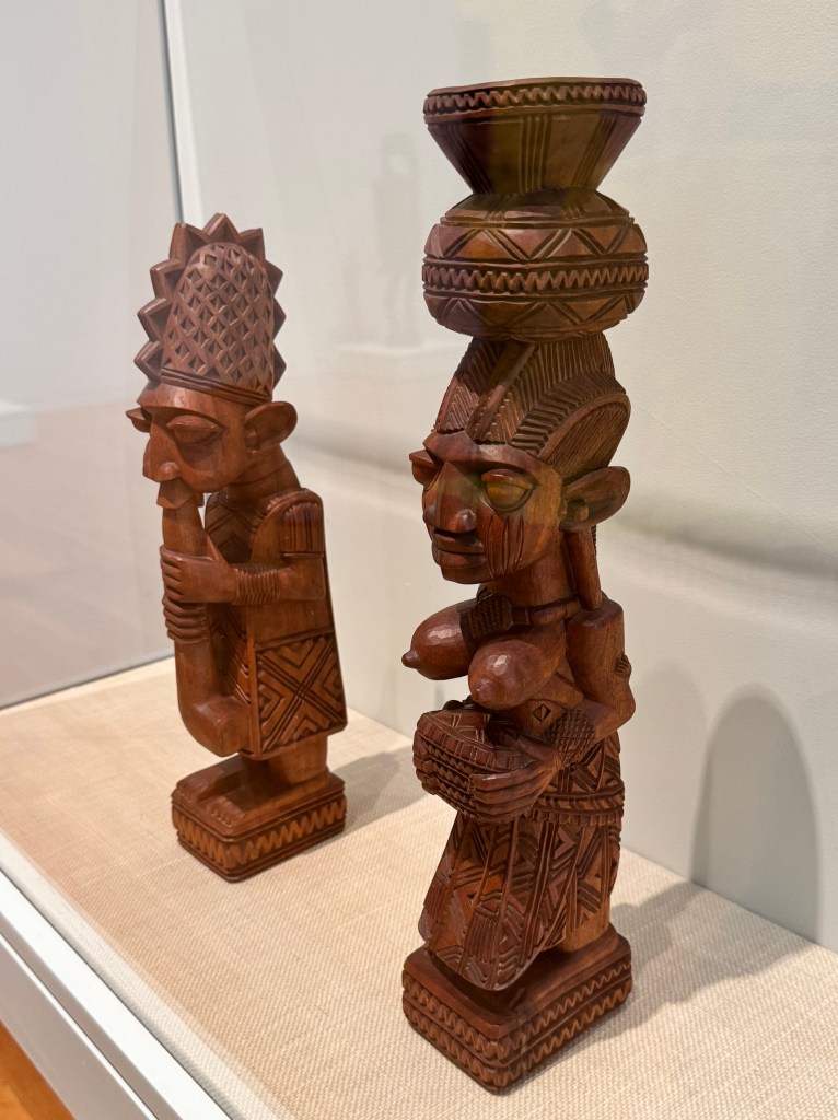 Two wooden sculptures from Nigeria; the following was on a plaque next to the sculptures: 

Lamidi Olonade Fakeye
Nigeria, 1928-2009
The Flute Player (left) and Shango's Wife (right)
Carved before 1967
Wood
Gift of Warren and Lynn Tacha, 2019/2.80 &.79
Provenance: Lynn Tacha 1967
These two figures were purchased directly from the artist Lamidi Fakeye by a Peace Corps volunteer stationed in Nigeria, right before she was evacuated from the country in 1967 following the outbreak of a brutal civil war. Fakeye was a fifth-generation carver who became a favorite among Peace Corps volunteers and other foreigners stationed in Nigeria.
His carvings gained such a reputation that he was asked to make them for buildings around the world, including the Kennedy Center in Washington, DC, and he was a frequent artist-in-residence at Western Michigan University and other universities in the United States and Europe. While Fakeye's carvings reference the Yoruba cultural traditions he was raised in, they were not meant to be used in ceremonies or rituals. His work was intended to be exported and displayed in collectors' homes and in museums.