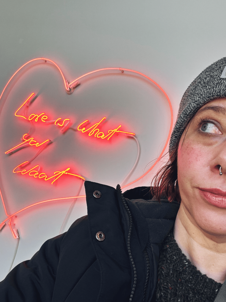 a short-haired woman is looking sideways at a neon sign that says "Love is what you want"